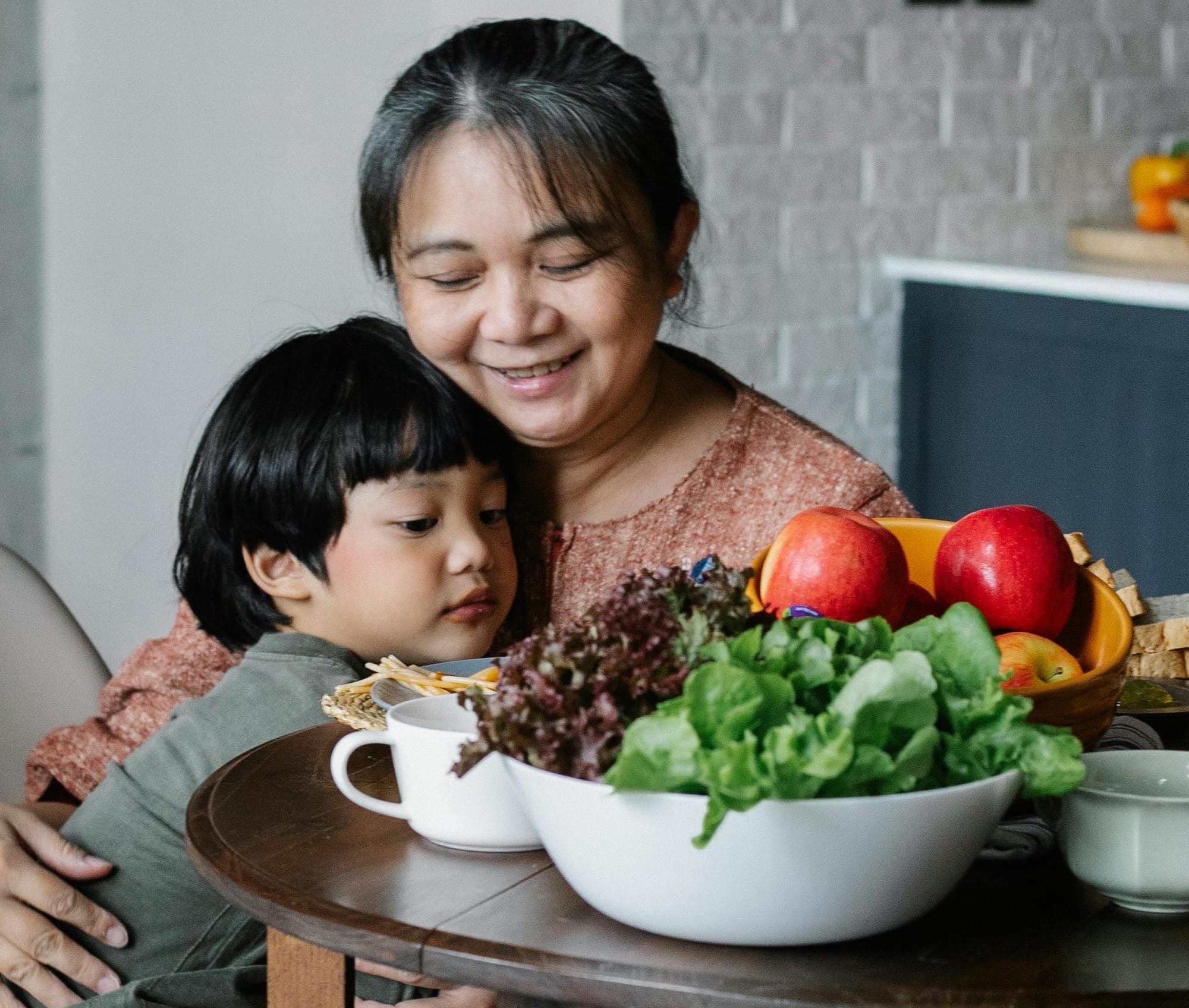 A smiling grandmother hugs her grandchild, who has a sober expression. Bowls of greens and fruit are on the table in front of them.