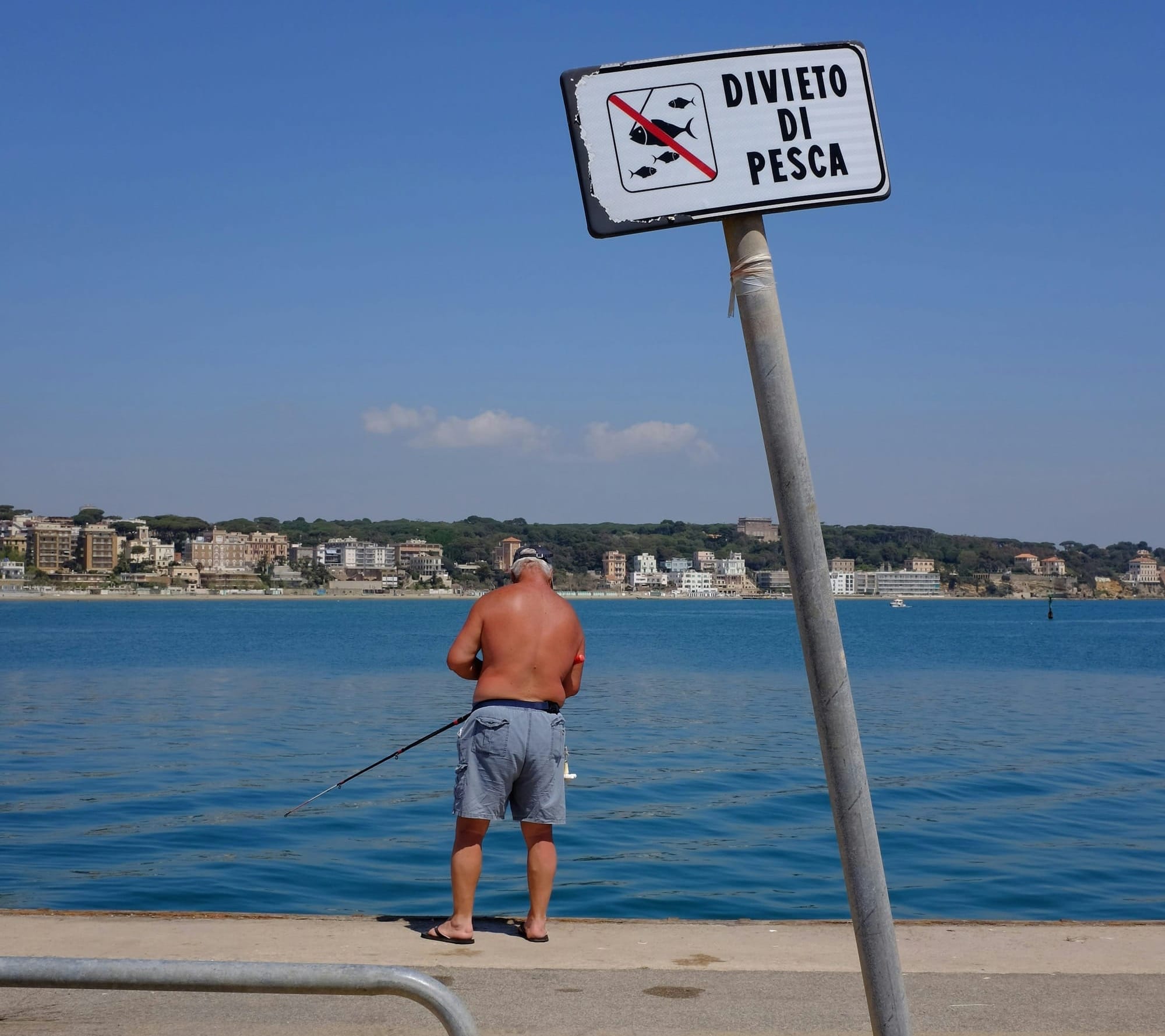 An older man fishes by a sign reading, "Divieto di pesca."