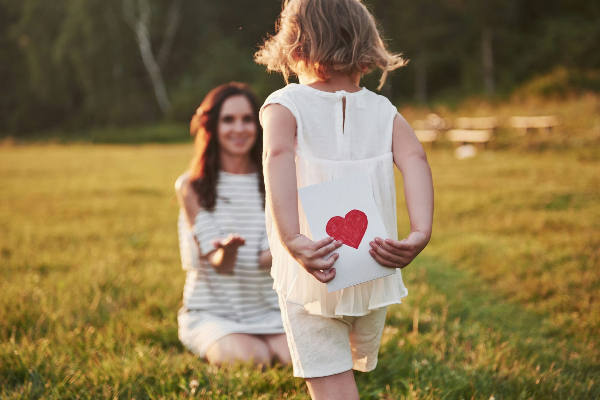 A child hides a card with a hand drawn heart behind her back. It looks as if she is about to give it to her mother, who is kneeling in the grass. 