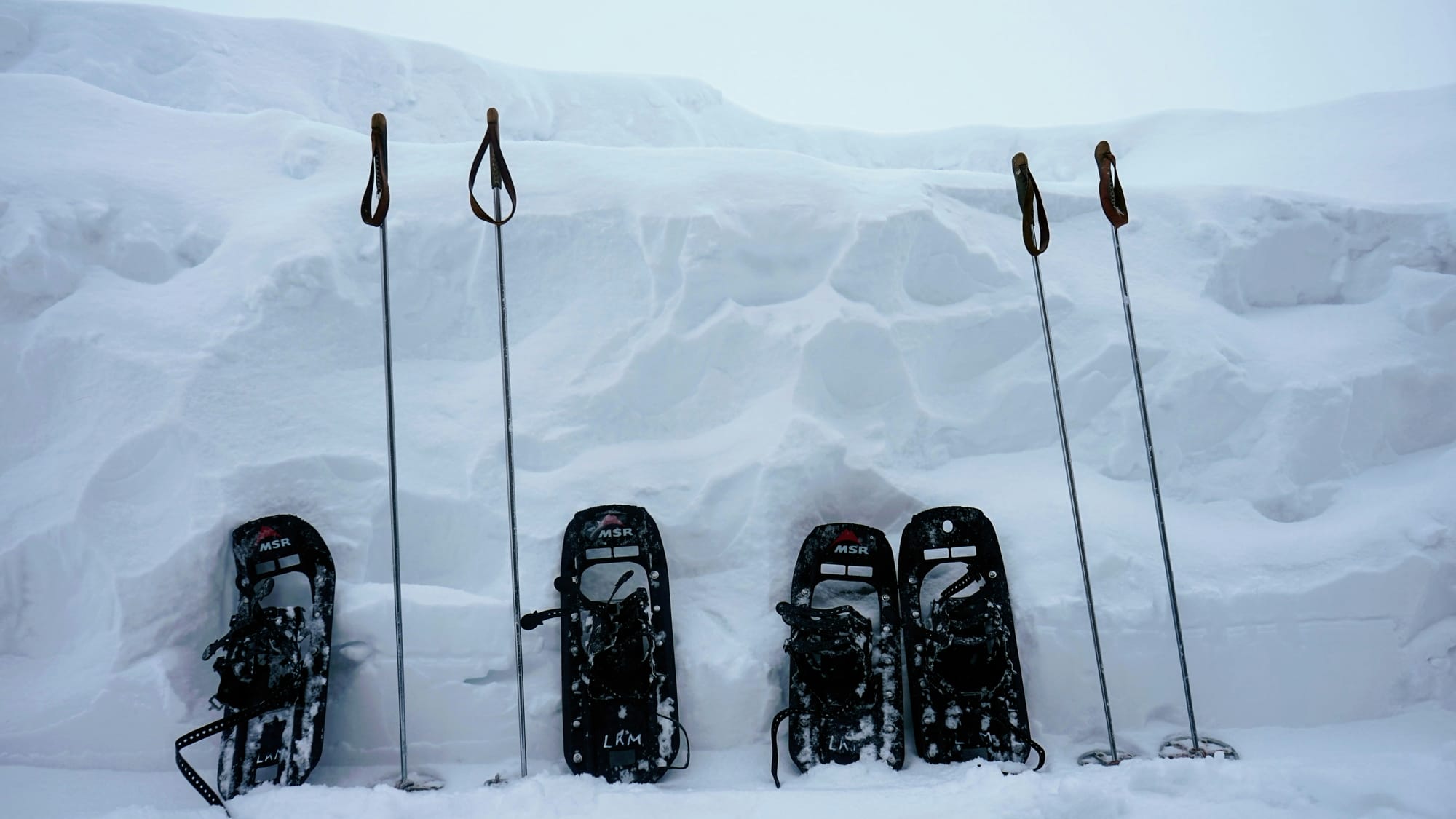 Four snowshoes and four poles lined up against a high snowbank.