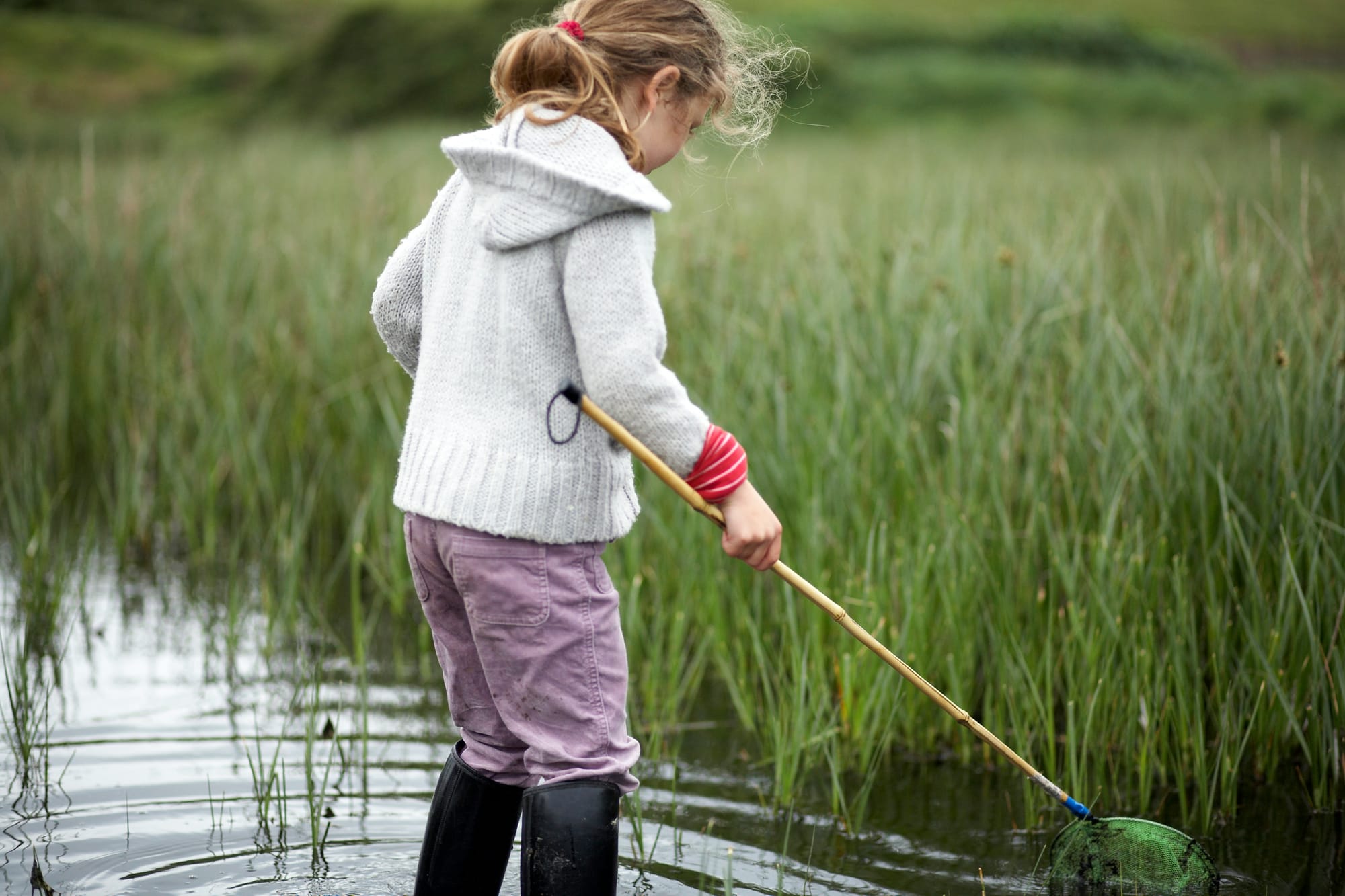 Photo by Getty Images for Unsplash.A girl wearing wading boots and carrying a small net walking in a grassy swamp.