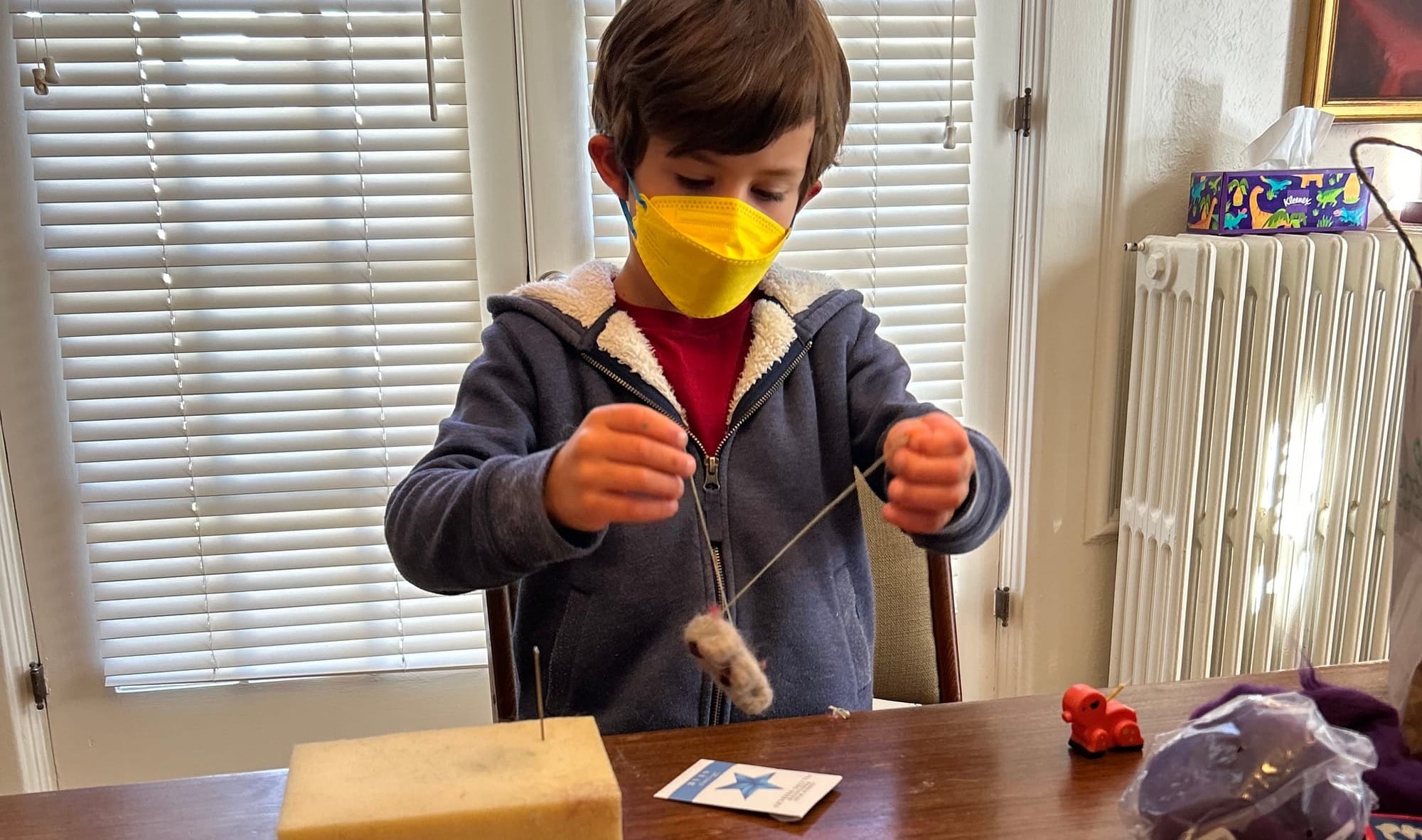 A child pulls a length of embroidery thread through his needle-felted sheep so he can hang it on a baby mobile.