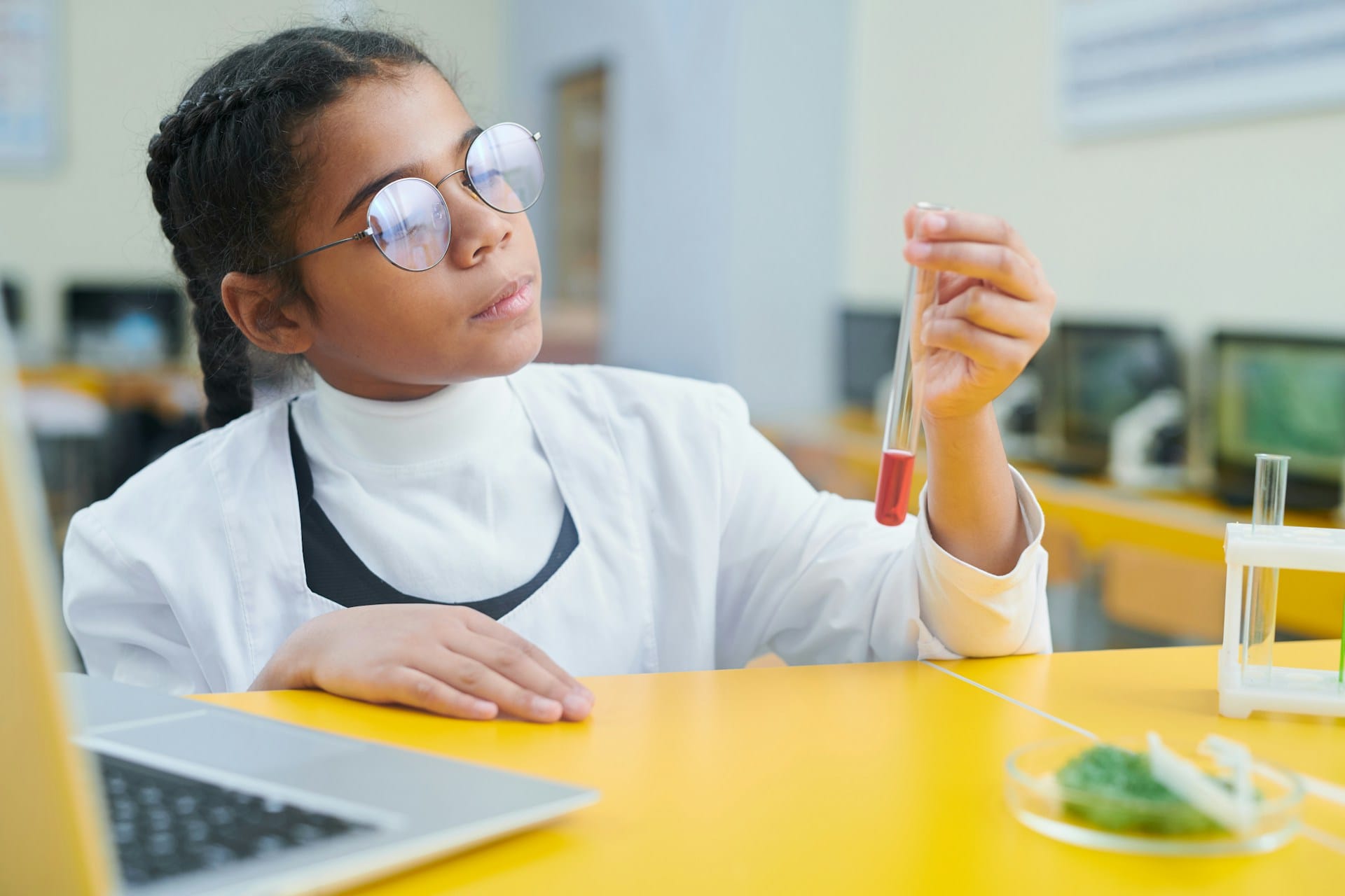 A girl examines a test tube containing an orange liquid. Laptop in the foreground. 
