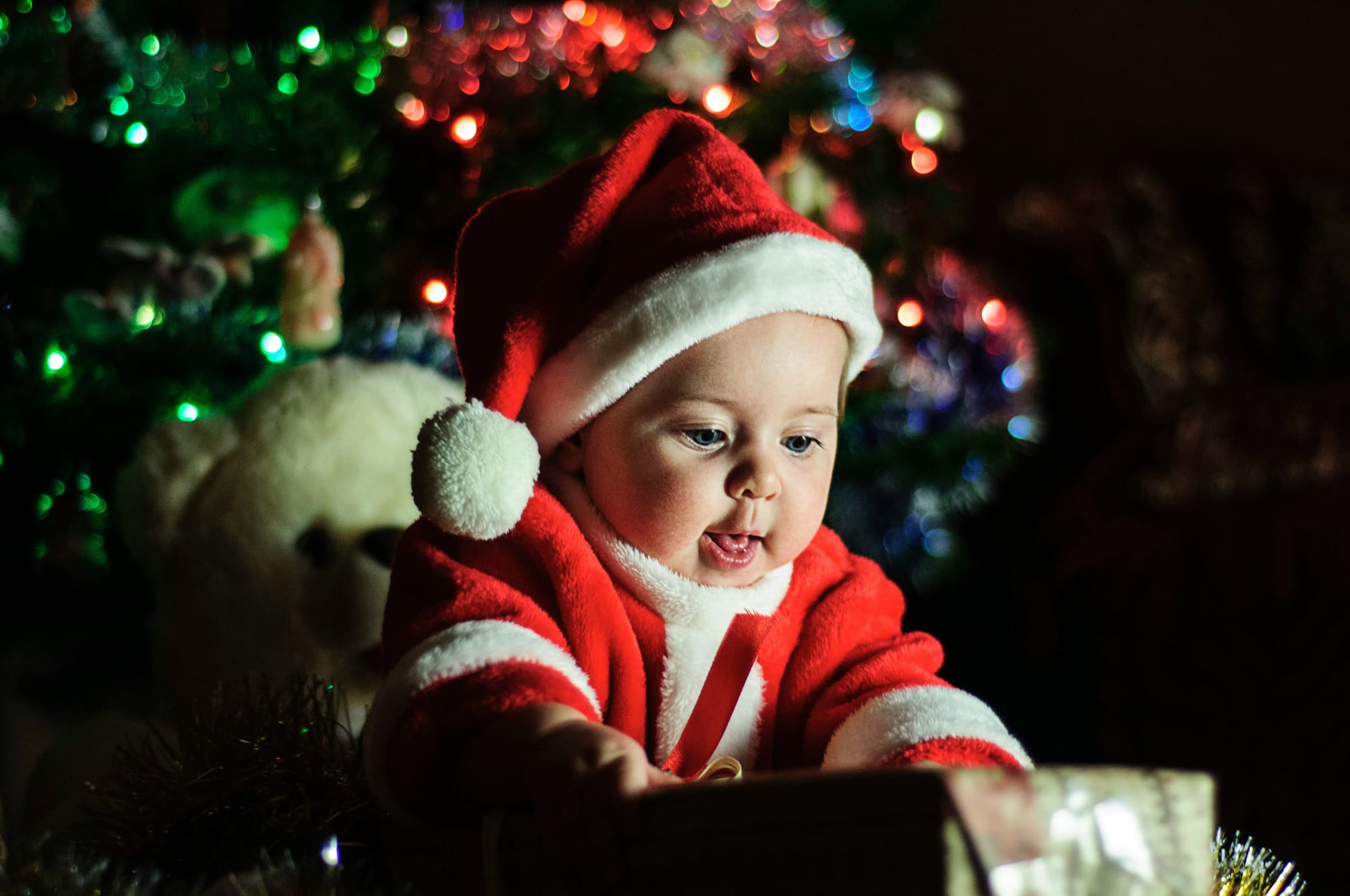 A baby in a Santa Claus suit, seated in front of a Christmas tree, seeming delighted with a wrapped gift.
