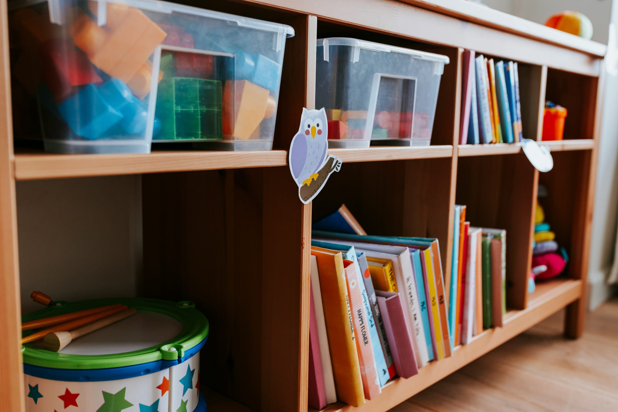A bookshelf contains bins of toys, a toy drum, books, and other toys.
