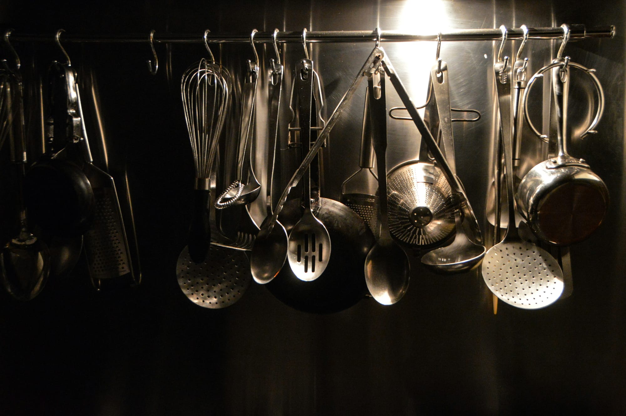 Spoons, ladles, whisks, pots, tongs, and other metal kitchen utensils hang from a rod.
