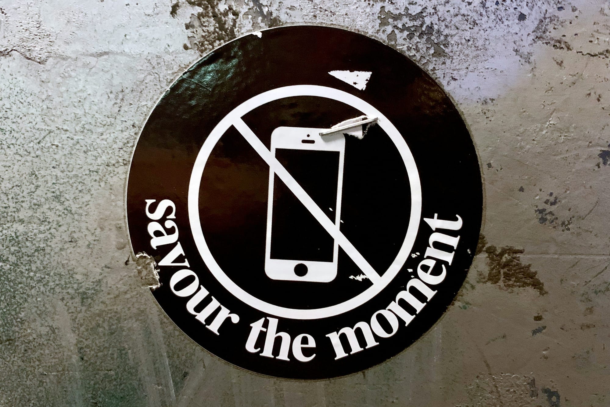 Circular black-and-white poster on a painted concrete wall, picturing a phone with the "no" sign over it, and the words, "Savour the moment."