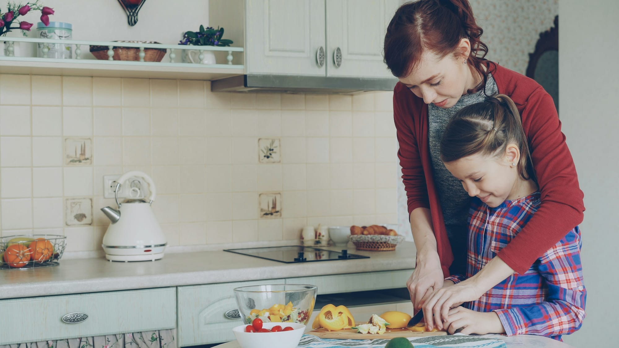 A mother shows her daughter how to slice a yellow pepper, the mother's arms embracing the girl and her hands guiding the girl's hands.