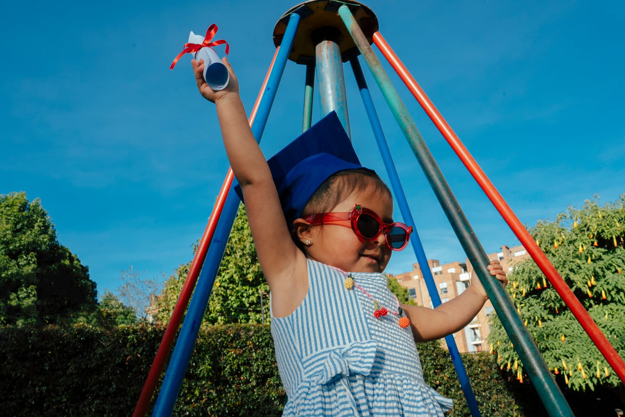 A preschooler in graduation cap and sunglasses excitedly raises her diploma.